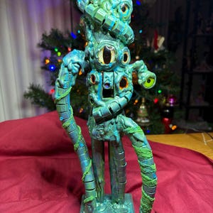 May include: A teal and green alien-like sculpture with multiple eyes and tentacle-like arms. The sculpture has a textured surface and is mounted on a square base. The eyes are yellow and brown.