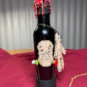 Puede incluir: Una botella de vidrio negro con una tapa de cera roja, adornada con clavos y una base de cuerda negra. Una etiqueta dice "Voodoo Venom Vial". Se adjunta una pequeña muñeca vudú y alfileres decorativos. La botella mide aproximadamente 25 cm de alto.