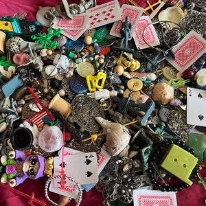 Op de afbeelding: Een verzameling van diverse snuisterijen en speelgoed, waaronder speelkaarten, speelgoed soldaten, kralen, knopen en een schelp. De items zijn verspreid over een rood stoffen oppervlak.