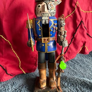 May include: A handcrafted nutcracker in a steampunk style. It features a blue coat with gold trim, a gray hat, and a mechanical arm. The nutcracker holds a sword and a staff with a glowing green orb.
