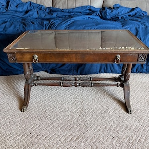 May include: A rectangular wooden coffee table with a glass top surface. The table features a dark brown finish, two metal ring pulls on the front drawer, carved floral details on the corners, and four legs with carved accents and claw feet.