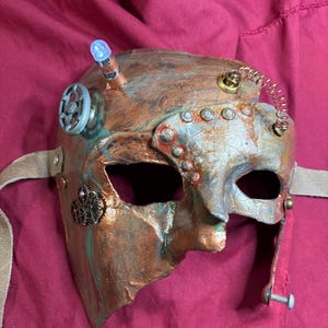 May include: A steampunk-style mask with a copper and silver finish. The mask features rivets, gears, and a small light. A leather strap is attached for wearing. The mask has an aged, weathered appearance.