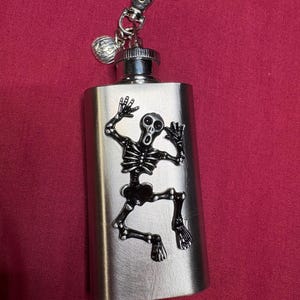 May include: A stainless steel flask with a black skeleton design. The flask has a silver cap, a small charm, and a clip for attaching to a bag or belt loop. The background is a solid maroon color.