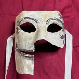 May include: A cream-colored masquerade mask with silver-colored decorative accents and eye holes. The mask has a broken edge on the right side and white ribbons attached for securing it. The mask is set against a red background.