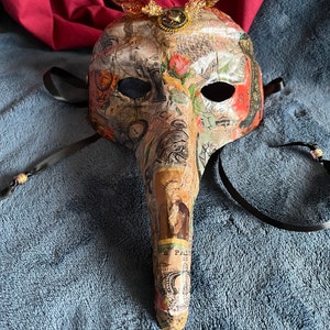 May include: A masquerade mask with a long, pointed nose, decorated with paper scraps, a clock face, and a butterfly with gold accents. The mask has black ribbon ties.