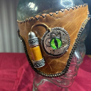 以下が含まれることがあります： 緑の目をした茶色の革製の steampunk マスク。銀と金の歯車と金と黄色のチューブが付いている。