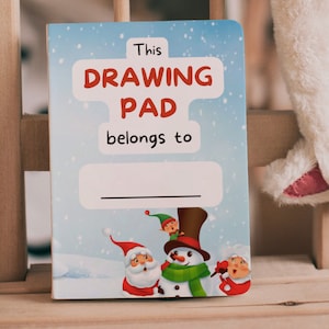 May include: A children's drawing pad with a winter theme. The cover reads "This DRAWING PAD belongs to" above a blank space for a name. Santa Claus, a snowman, and an elf are illustrated at the bottom. The background is a light blue with falling snowflakes.