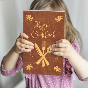 May include: A brown "Magic Cookbook" with gold lettering and illustrations of a knife, fork, and spoon. The cover has decorative flourishes and star accents. The cookbook is held by a person wearing a pink dress.