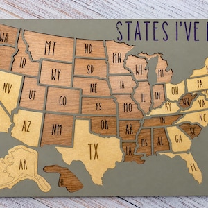 May include: Wooden United States map with individual states in varying shades of brown and tan. The text "STATES I'VE RUN" is written across the top in dark purple. The map is mounted on a gray background.