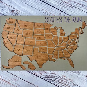 May include: A rectangular wooden wall plaque featuring a laser-cut map of the United States with individual state borders and abbreviations. The text 'STATES I'VE RUN' is printed in purple in the upper right corner against a light gray background.