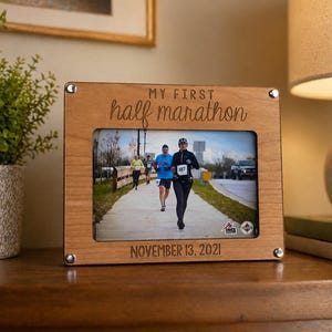 May include: Wooden photo frame with the text "MY FIRST half marathon" and "NOVEMBER 13, 2021". The frame holds a photo of runners in a race. The frame is on a wooden surface.