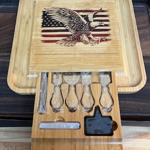 May include: A wooden cheese board with a drawer that holds a set of cheese knives. The board is decorated with an American flag and an eagle.