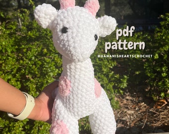 May include: White crocheted giraffe amigurumi with pink ears and spots. The giraffe is being held by a hand. The text "pdf pattern @HANANISHEARTSCROCHET" is visible in the image.