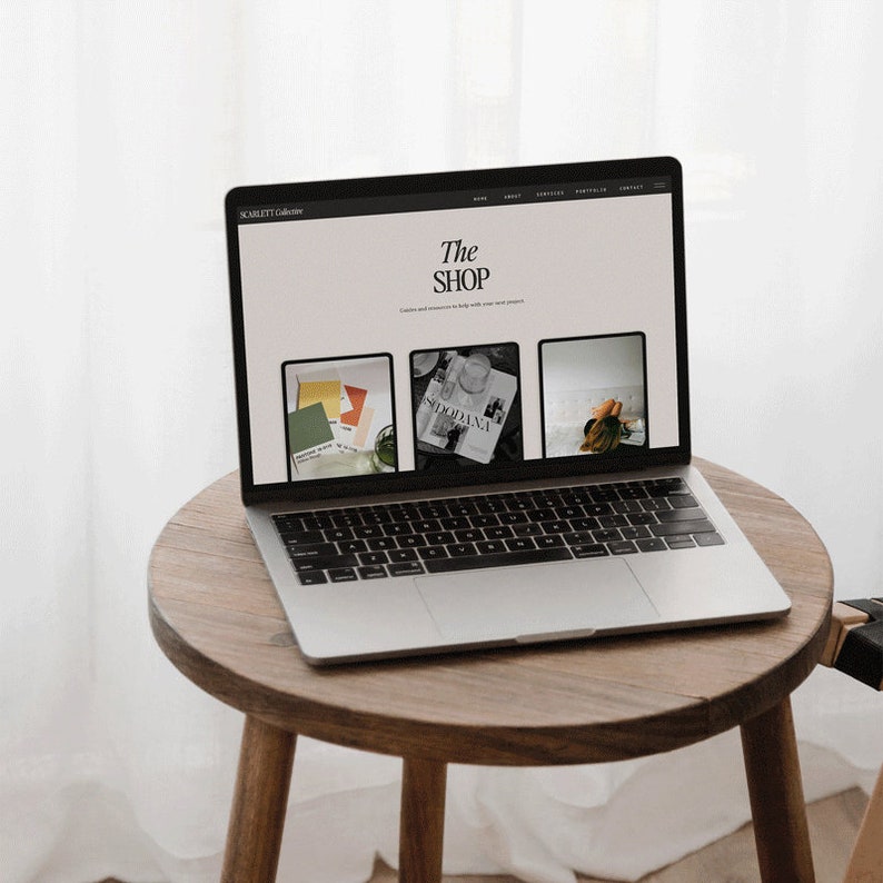 May include: A laptop computer open to a website with the text "The Shop" on the screen. The website features images of a magazine, a set of paint swatches, and a person lying on a bed. The laptop is sitting on a wooden table.