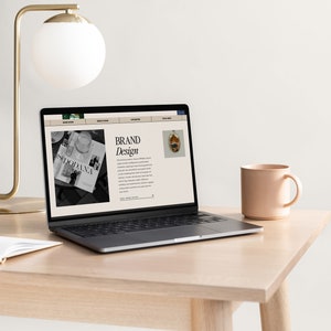 May include: A laptop computer with a website open on the screen. The website is about brand design and features a black and white photo of a magazine and a photo of a small, colorful object. The laptop is on a light wood desk with a white mug and a lamp.