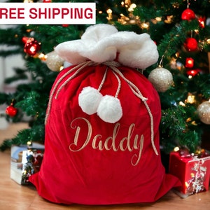 May include: Red velvet Santa sack with white faux fur trim and a gold embroidered "Daddy" on the front. The sack is sitting in front of a Christmas tree with ornaments.