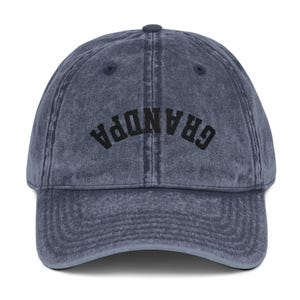 May include: A denim blue baseball cap with the word "GRANDPA" embroidered in black across the front. The cap has a curved brim and a button on top. The fabric has a vintage, worn look.