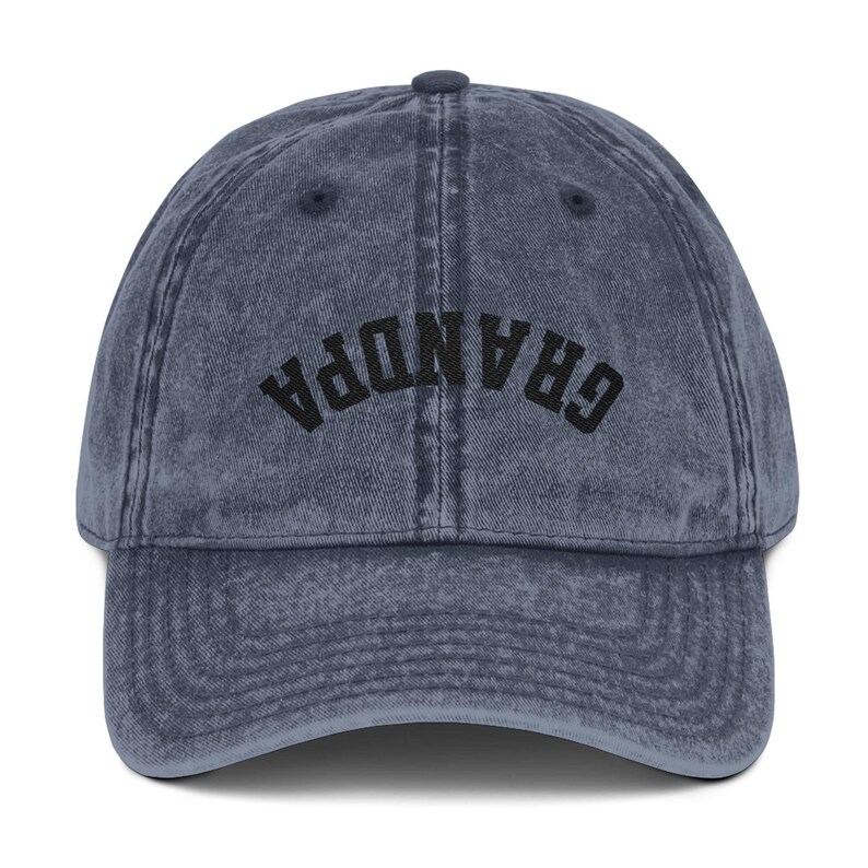 May include: A denim blue baseball cap with the word "GRANDPA" embroidered in black across the front. The cap has a curved brim and a button on top. The fabric has a vintage, worn look.