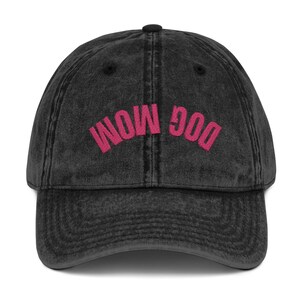 May include: A black, vintage-style baseball cap with the embroidered text "DOG MOM" in bright pink. The cap has a curved brim and a button on top. The fabric appears worn, giving it a retro look.