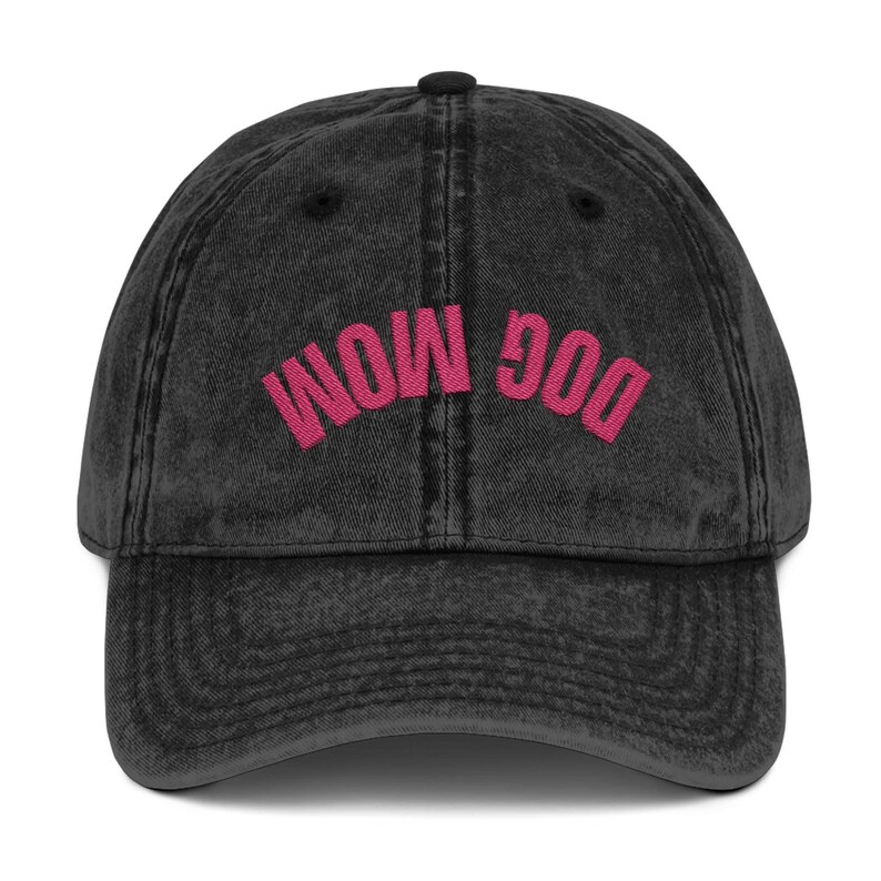 May include: A black, vintage-style baseball cap with the embroidered text "DOG MOM" in bright pink. The cap has a curved brim and a button on top. The fabric appears worn, giving it a retro look.