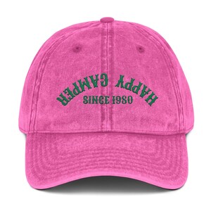 May include: A pink baseball cap with the words "HAPPY CAMPER" embroidered in green across the front, with "SINCE 1980" underneath. The cap has a curved brim and a fabric strap closure.