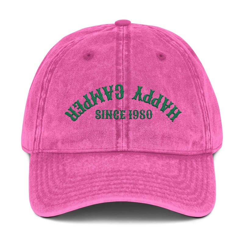 May include: A pink baseball cap with the words "HAPPY CAMPER" embroidered in green across the front, with "SINCE 1980" underneath. The cap has a curved brim and a fabric strap closure.