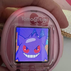 Puede incluir: Mascota virtual Tamagotchi rosa con un personaje Gengar morado en la pantalla. El dispositivo tiene una carcasa rosa con el texto "Tamagotchi" y "Sanrio characters". Se ve un bot&oacute;n rojo debajo de la pantalla.