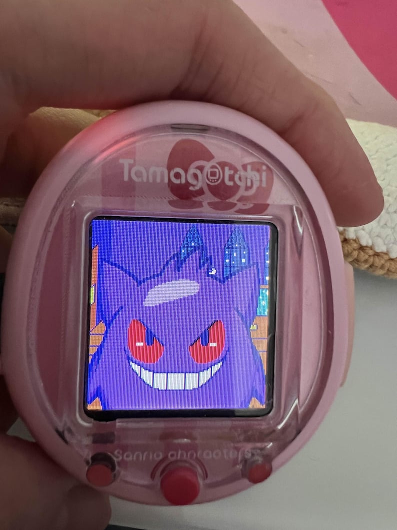 Puede incluir: Mascota virtual Tamagotchi rosa con un personaje Gengar morado en la pantalla. El dispositivo tiene una carcasa rosa con el texto "Tamagotchi" y "Sanrio characters". Se ve un bot&oacute;n rojo debajo de la pantalla.