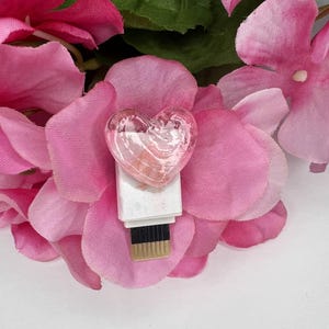 Puede incluir: Una unidad USB transparente en forma de coraz&oacute;n se encuentra sobre una base blanca, rodeada de p&eacute;talos de flores rosas y hojas verdes. La unidad USB tiene un tono rosado y est&aacute; colocada en el centro de la imagen. El fondo es blanco.