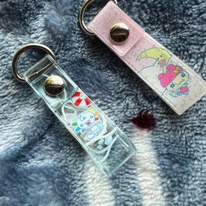 May include: Two rectangular keychains with silver metal rings and snap buttons. One is clear with a colorful axolotl graphic and a red and white life preserver. The other is light pink with glitter and a graphic of a character with a yellow crescent moon.