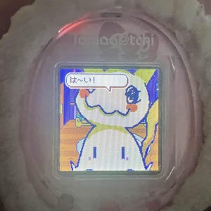 Puede incluir: Una mascota digital Tamagotchi rosa con una funda de felpa blanca. La pantalla muestra un personaje de dibujos animados con una burbuja de di&aacute;logo que contiene texto japon&eacute;s. El dispositivo est&aacute; dise&ntilde;ado para el cuidado e interacci&oacute;n de mascotas virtuales.