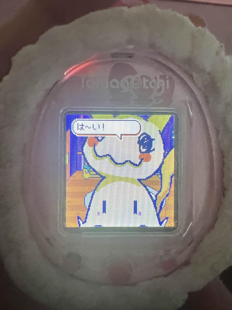 Puede incluir: Una mascota digital Tamagotchi rosa con una funda de felpa blanca. La pantalla muestra un personaje de dibujos animados con una burbuja de di&aacute;logo que contiene texto japon&eacute;s. El dispositivo est&aacute; dise&ntilde;ado para el cuidado e interacci&oacute;n de mascotas virtuales.