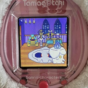 Puede incluir: Un dispositivo de mascota virtual Tamagotchi rosa con una pantalla que muestra una escena de dibujos animados. El dispositivo tiene un borde blanco y esponjoso y botones rojos. Los textos "Tamagotchi" y "Sanrio characters" son visibles.