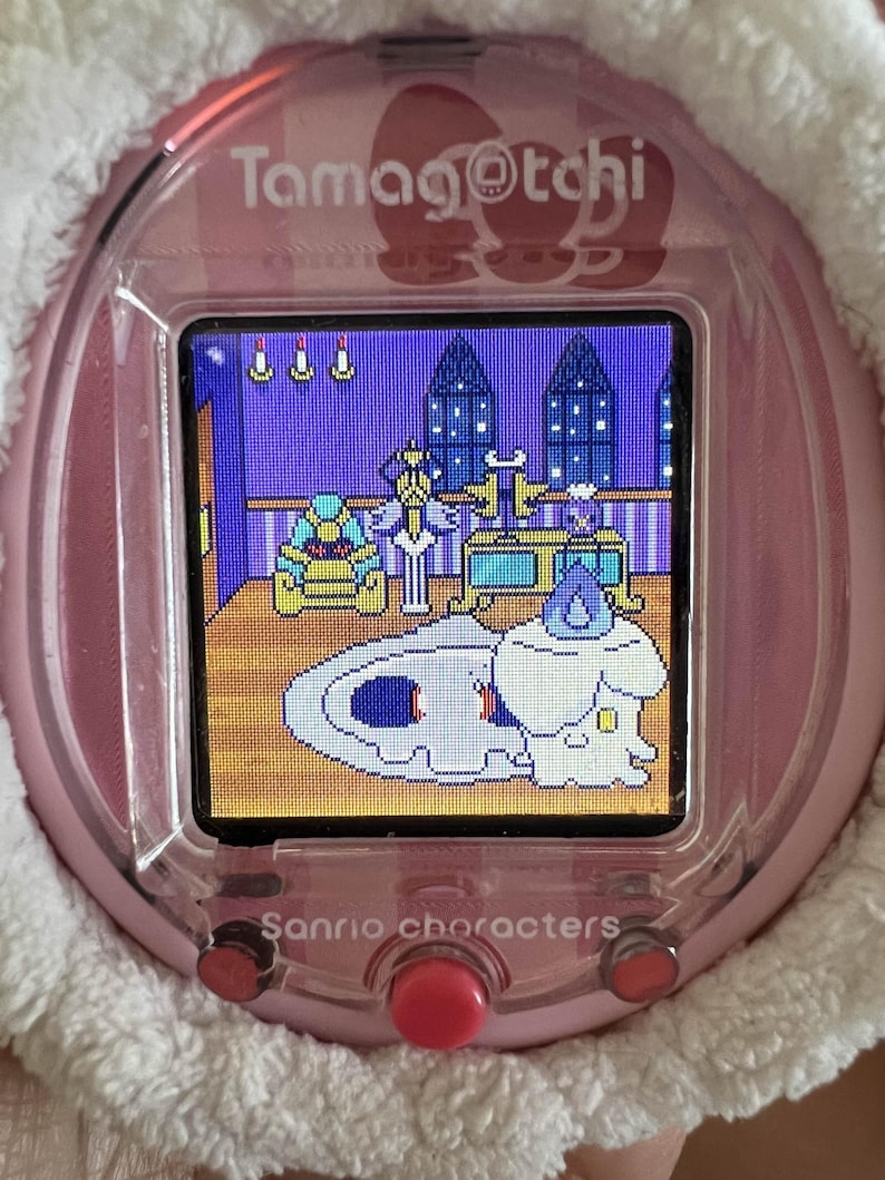 Puede incluir: Un dispositivo de mascota virtual Tamagotchi rosa con una pantalla que muestra una escena de dibujos animados. El dispositivo tiene un borde blanco y esponjoso y botones rojos. Los textos "Tamagotchi" y "Sanrio characters" son visibles.