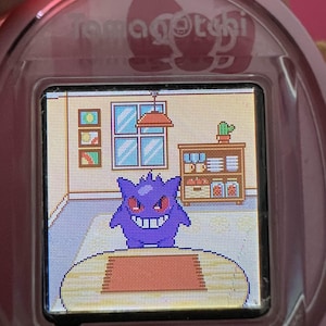 Puede incluir: Dispositivo Tamagotchi rosa con un personaje Gengar pixelado en un entorno de habitaci&oacute;n. La pantalla muestra un Gengar morado con ojos rojos y una amplia sonrisa. El texto "Sanrio characters" est&aacute; en la parte inferior.