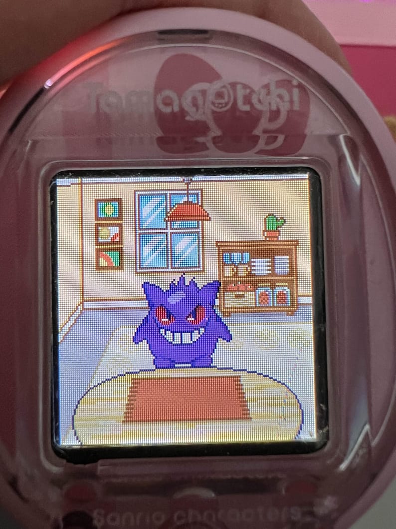 Puede incluir: Dispositivo Tamagotchi rosa con un personaje Gengar pixelado en un entorno de habitaci&oacute;n. La pantalla muestra un Gengar morado con ojos rojos y una amplia sonrisa. El texto "Sanrio characters" est&aacute; en la parte inferior.