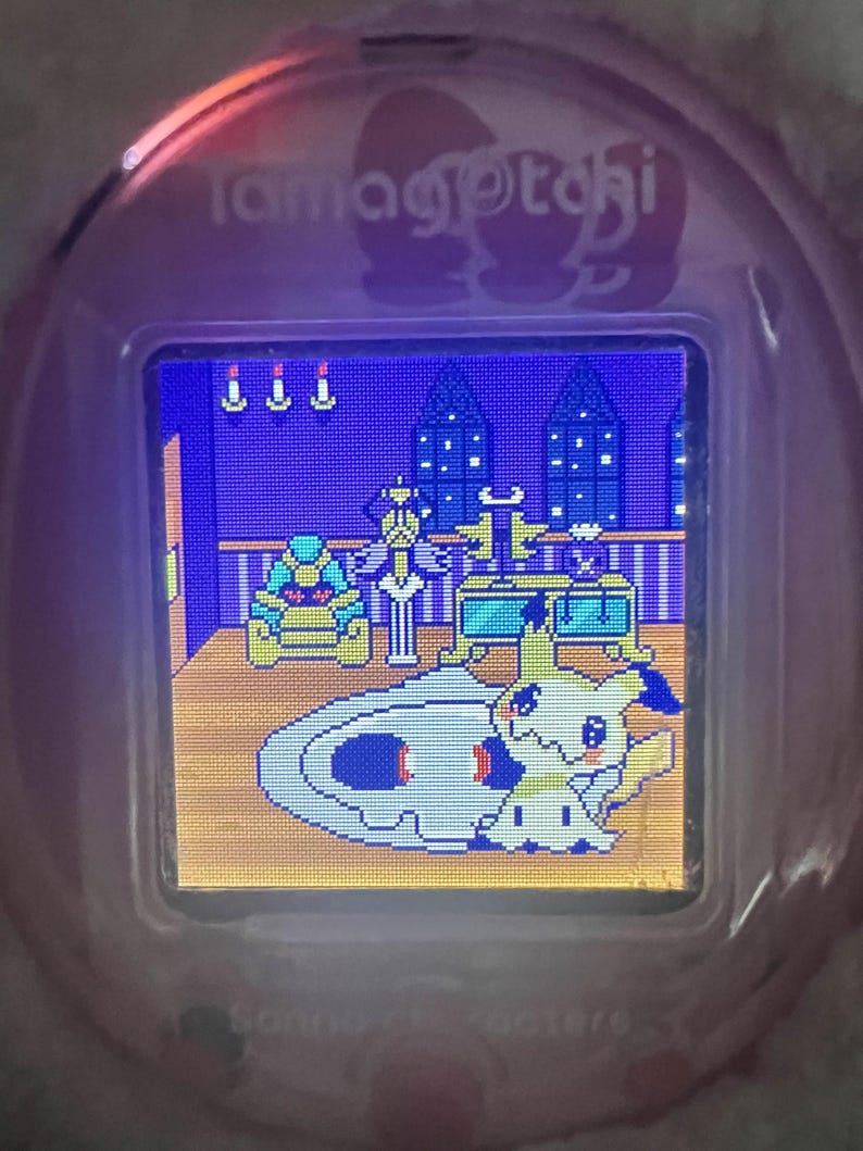 Puede incluir: Un dispositivo Tamagotchi rosa con una pantalla digital que muestra una escena pixelada. La escena presenta un personaje de dibujos animados, muebles y una alfombra estampada. El dispositivo tiene la palabra "Tamagotchi" impresa en la parte superior.