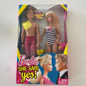 May include: Barbie She Said Yes! doll set featuring a blonde doll in a black and white swimsuit and a doll with brown hair in a red shirt and pink shorts. The dolls are holding hands and the box has a pink heart with the words "Together Again" on it.
