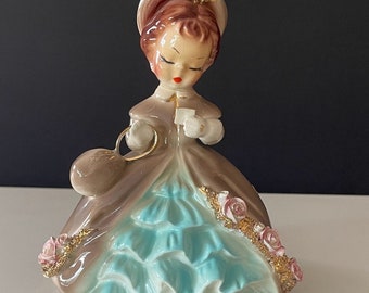 Peut inclure: Une figurine en porcelaine d'une femme vêtue d'une robe brune et bleue avec un chapeau blanc et des fleurs roses. Elle tient une petite enveloppe blanche dans sa main.