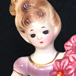 May include: A porcelain figurine of a girl with brown hair, wearing a lavender dress with gold trim, holding a bouquet of pink flowers.