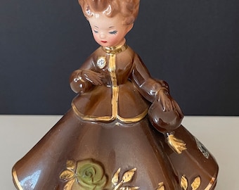 Może przedstawiać: Vintage ceramiczna figurka kobiety w brązowej sukni ze złotymi wykończeniami. Figurka ma zieloną kokardę i zielone róże na spódnicy. Kobieta trzyma małą torebkę. Figurka ma około 15 cm wysokości.
