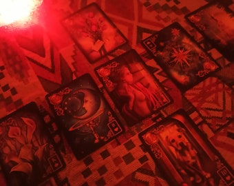 Puede incluir: Un despliegue de lectura de cartas de tarot con una vela roja en el fondo. Las cartas están dispuestas sobre una tela estampada. Las cartas muestran imágenes de una luna, una mujer, un perro y una brújula.