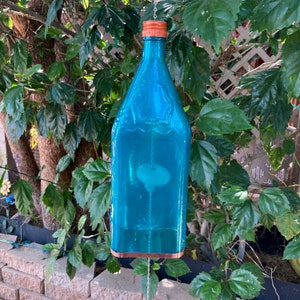 May include: A teal blue glass bottle wind chime with a copper top and a white disc inside. The wind chime is hanging from a chain.