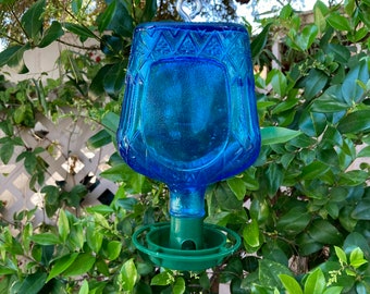 May include: A blue glass hummingbird feeder with a green plastic base and a metal hook. The feeder is hanging from a tree branch.