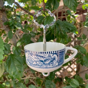 May include: A white teacup with a blue and white illustration of a horse-drawn carriage. The teacup is suspended by a silver metal handle with an ornate design. The teacup is hanging from a tree branch.
