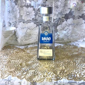 May include: A clear glass bottle of 1800 Tequila Blanco with a silver cap and a blue label. The label features the brand name and the word "Blanco". The bottle is on a gold-colored, floral-patterned surface.