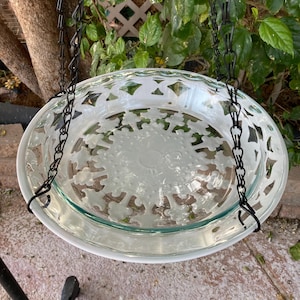 May include: A clear glass bird bath with a white rim and decorative cut-out design. The bird bath is suspended by a black chain, creating a hanging effect. The design features a series of floral shapes. The bird bath is outdoors.