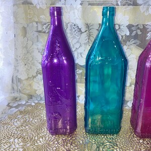 May include: Three glass bottles with embossed designs. The bottles are purple, teal, and pink. The bottles are sitting on a gold-colored surface.