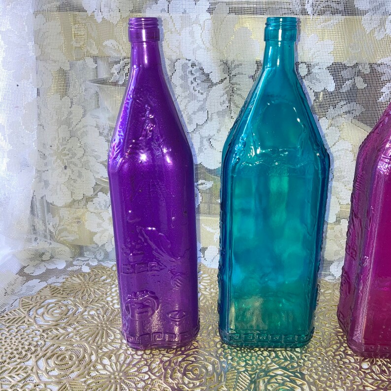 May include: Three glass bottles with embossed designs. The bottles are purple, teal, and pink. The bottles are sitting on a gold-colored surface.