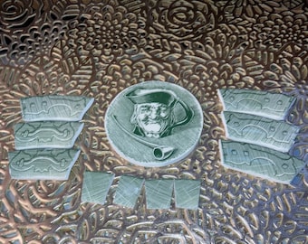 May include: A set of green and white ceramic tiles with a design of a man with a pipe. The tiles are arranged in a circle around a central tile with a portrait of the man.